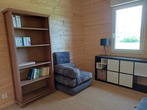 A room with a wooden bookshelf, a patterned armchair, a lamp, and a window with a view.