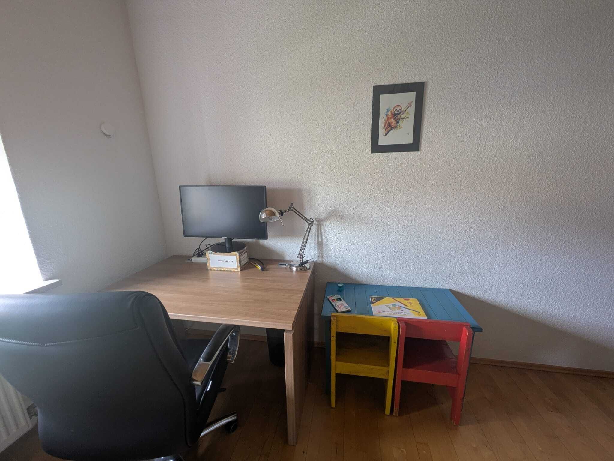 A room with a desk, a computer, a chair, a small table with a chair, and a framed picture on the wall.