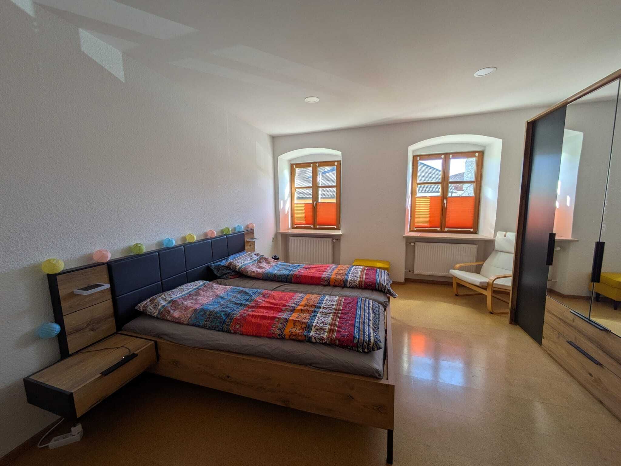 Ein Schlafzimmer mit einem Bett, zwei Fenstern, einem Sessel und einem Spiegel.