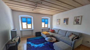 Una sala de estar con un sofá, un reposapiés, una mesa de centro y un tapete azul estampado. Hay dos ventanas con persianas azules, un radiador y un ventilador de techo.