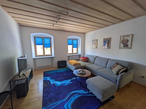 Un soggiorno con divano, poggiapiedi, tavolino e un tappeto azzurro decorato. Sono presenti due finestre con persiane blu, un termosifone e una ventola a soffitto.