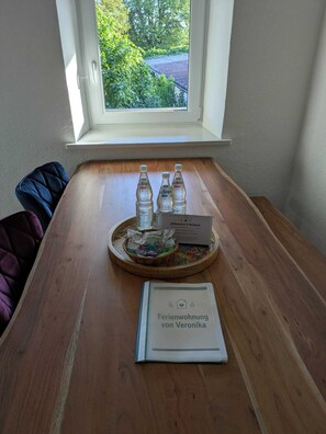 Un tavolo di legno con un cartello di benvenuto, tre bottiglie d'acqua e un libretto intitolato 'Ferienwohnung von Veronica' davanti a una finestra che si affaccia sul verde.