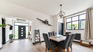 A modern dining area with a wooden table and grey upholstered chairs, a chandelier, and a view of greenery through a large window. - Stunning Retreat in Matlock Bath (Matlock)