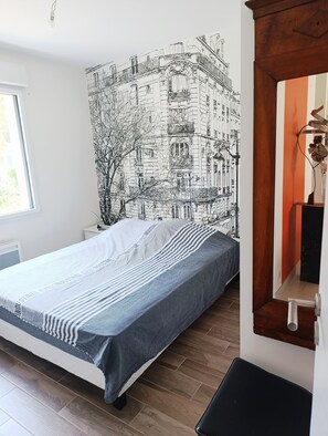 A bed with a striped bedspread, a wall with a black and white cityscape mural, a window, and a wooden floor.