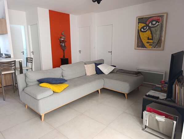 Un salon avec un canapé gris, un coin repas avec une table et des chaises blanches, et un radiateur.