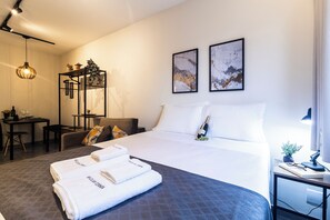 Quarto de hotel com cama, escrivaninha, cadeira e pequena mesa com uma planta e uma garrafa.