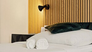 Ein ordentlich bezogenes Bett mit weißen Leinen und aufgerollten Handtüchern, ein dunkelgrünes Kopfkissen und eine an der Wand befestigte Lampe.