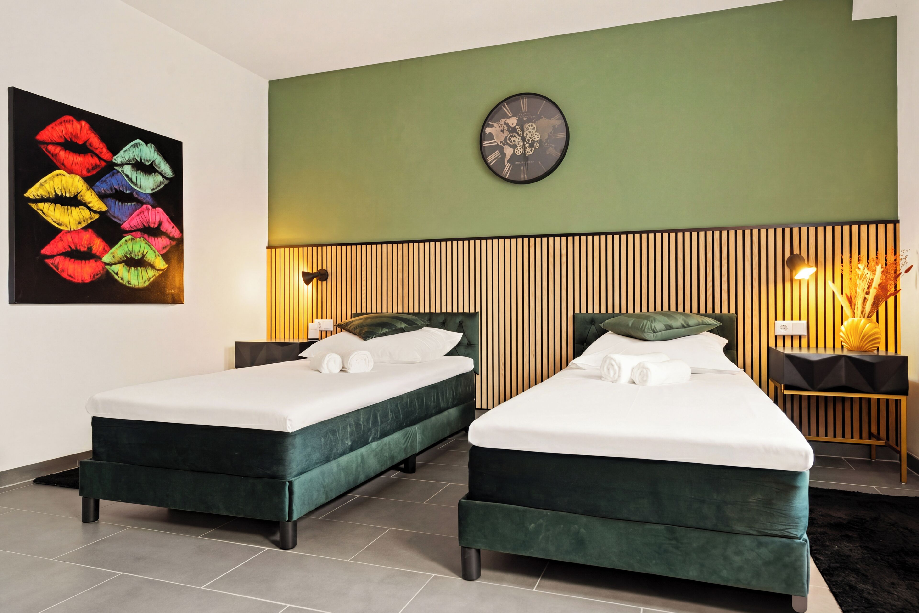 Camera d'albergo con due letti, una parete verde, una sveglia, un'opera d'arte con labbra e un comodino con una lampada.
