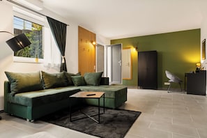 A modern living room with a sectional sofa, a small table, and a view of the outdoors through a window. - Modo Apartments Erkelenz | Pool and sauna (Erkelenz)