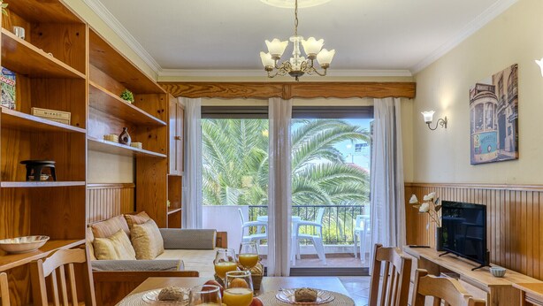 A dining area with a wooden table set for breakfast, a sofa, and a TV on a wooden stand. - #366 Cozy 1BR Retreat | Private Balcony (Albufeira)