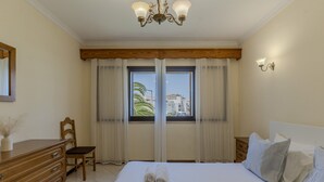 A hotel room with a large bed, a wooden dresser, a chair, a nightstand, a wall-mounted lamp, and a window with curtains. - #366 Cozy 1BR Retreat | Private Balcony (Albufeira)