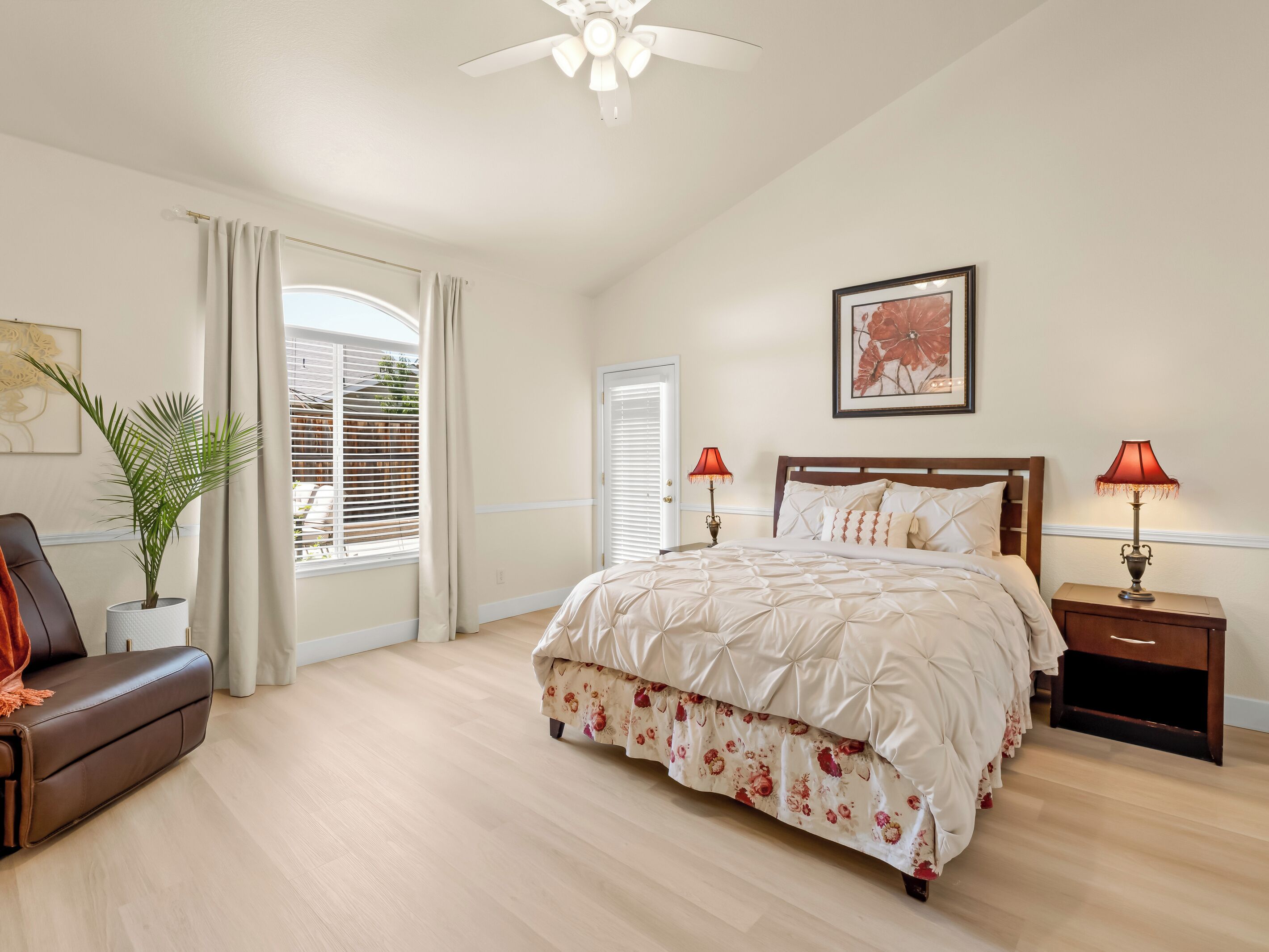 Quarto com cama, mesa de cabeceira, luminária, cadeira, planta em vaso e janela com cortinas.