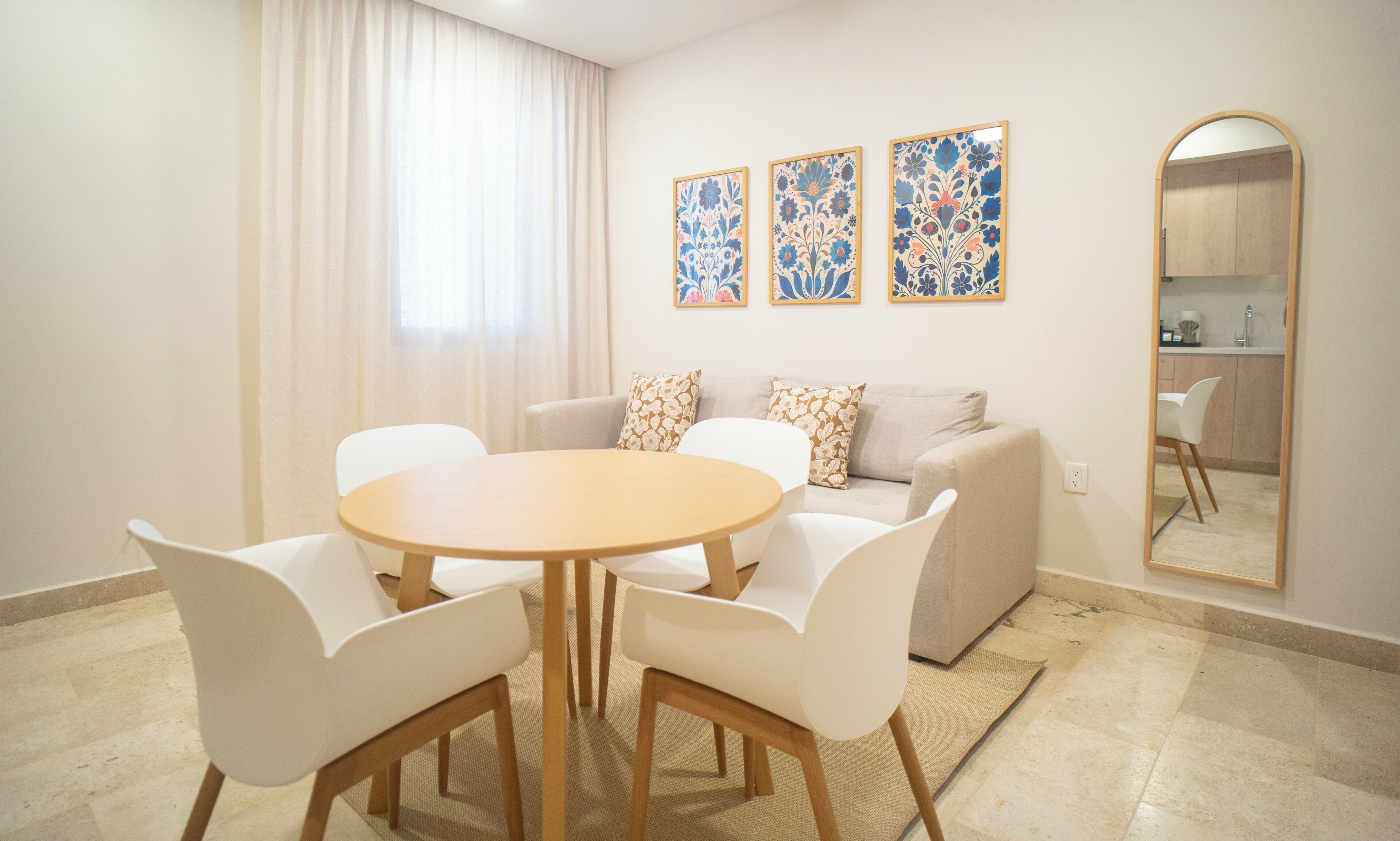 Uma sala de estar moderna com uma mesa redonda de madeira, cadeiras brancas, um sofá bege com almofadas florais, e uma cozinha visível através de um arco.