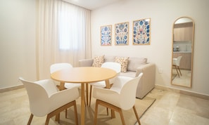 Una sala de estar moderna con una mesa redonda de madera, sillas blancas, un sofá beige con cojines estampados y una cocina visible a través de un arco.