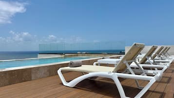 Zona de piscina con sillas de descanso, una barrera de cristal y vista al océano.