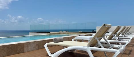 A poolside area with lounge chairs, a glass barrier, and a view of the ocean.