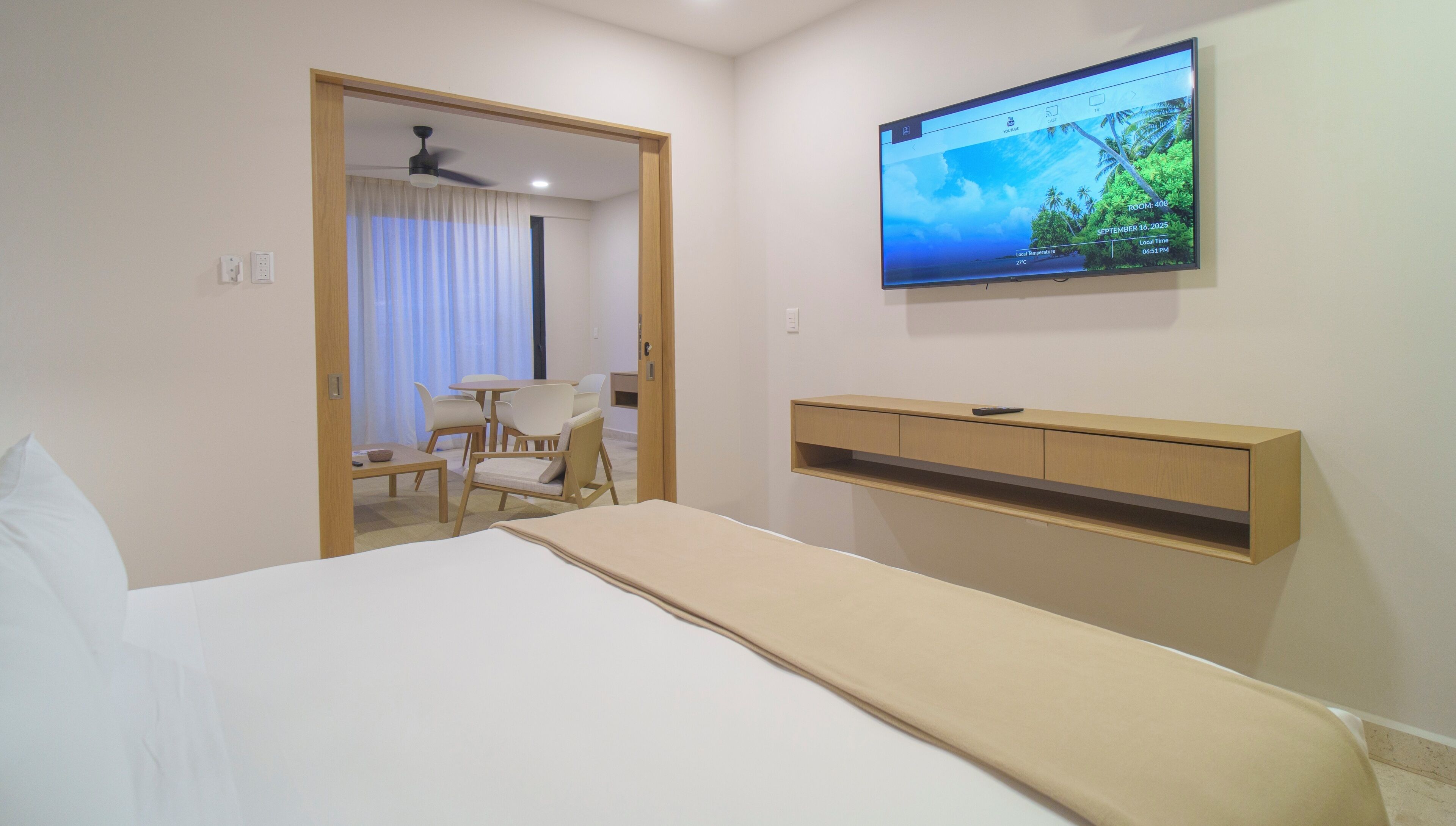 Habitación de hotel con una cama, televisor montado en la pared, escritorio con sillas y ventilador de techo.