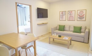 A modern living room with a sofa, a coffee table, a TV mounted on the wall, and a dining area with chairs and a table.