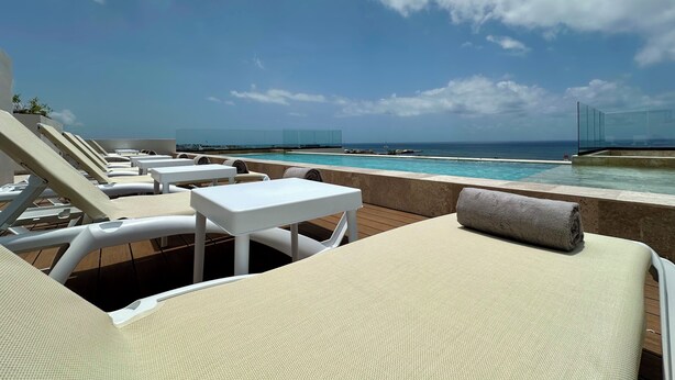 Un área junto a la piscina con sillones de descanso y mesas, vista despejada al océano, y una barrera de vidrio que separa la piscina de la playa.