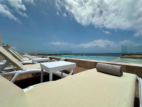 Zona de piscina amb gandules i taules, vista clara de l'oceà i una barrera de vidre que separa la piscina de la platja.
