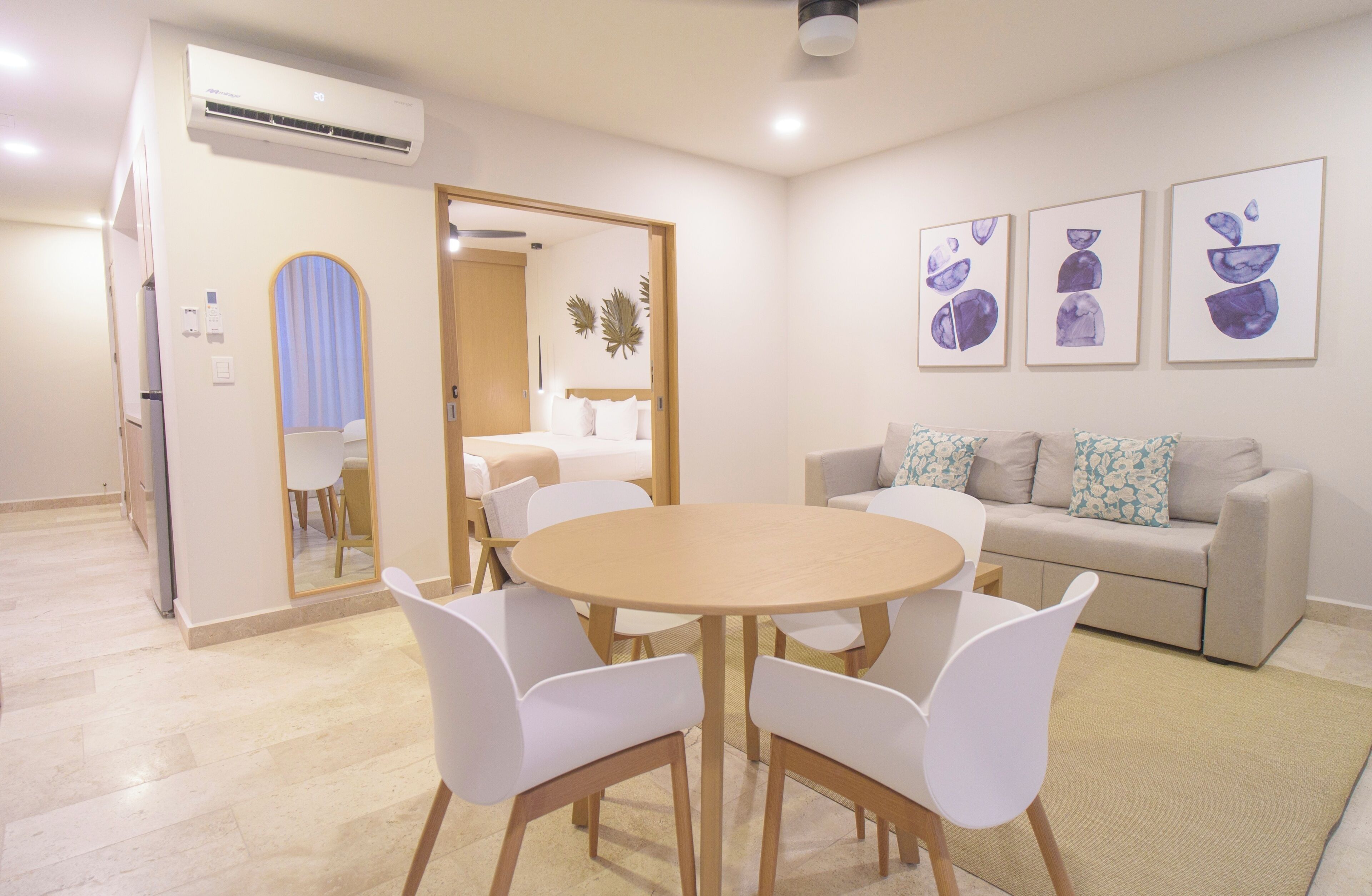 Una sala de estar moderna con una mesa redonda de madera y sillas blancas, un sofá color crema con cojines estampados y tres obras de arte enmarcadas en la pared.