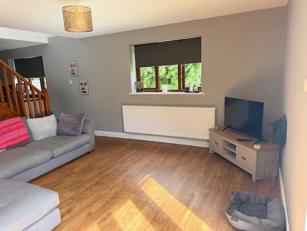 A living room with a grey sofa, a TV, a radiator, a window with a view, and a wooden staircase.