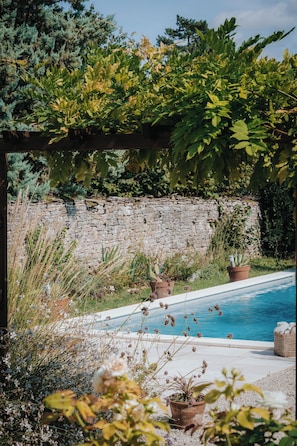 A pool area with a stone wall, potted plants, and a pergola with greenery.