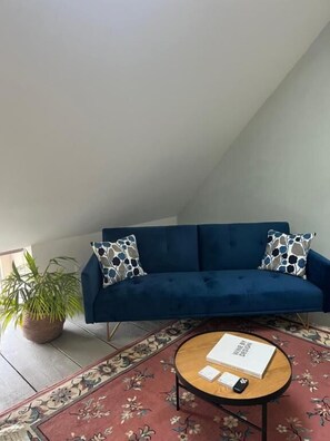 A room with a blue sofa, two patterned pillows, a round coffee table with a book and remote control, and a floral rug.