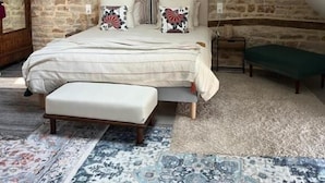 A bedroom with a stone wall, a bed with patterned pillows, a bench, and a large rug. - Maison Tassigny (Meursault)