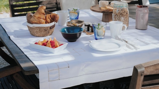 An outdoor dining setup with a table set for two, featuring a basket of croissants, a bowl of fruit, and various condiments. - Maison Tassigny (Meursault)
