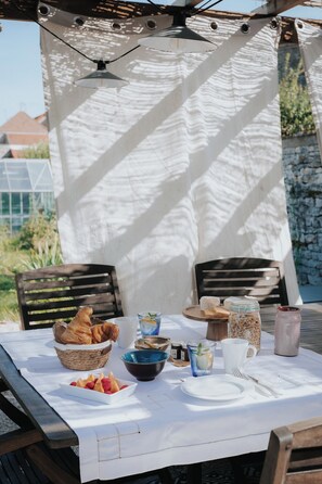 An outdoor dining setup with a table set for two, featuring a basket of croissants, a bowl of fruit, and various condiments. - Maison Tassigny (Meursault)