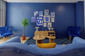 A room with a blue wall, two blue armchairs, a sofa with a yellow pillow, a wooden cabinet, and a potted plant.