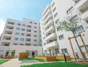 A multi-story residential building with balconies, a small garden area with wooden planters, and a clear sky.