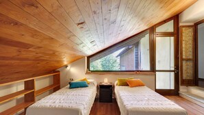 A room with two beds, wooden ceiling, large window, and a small table with a lamp.
