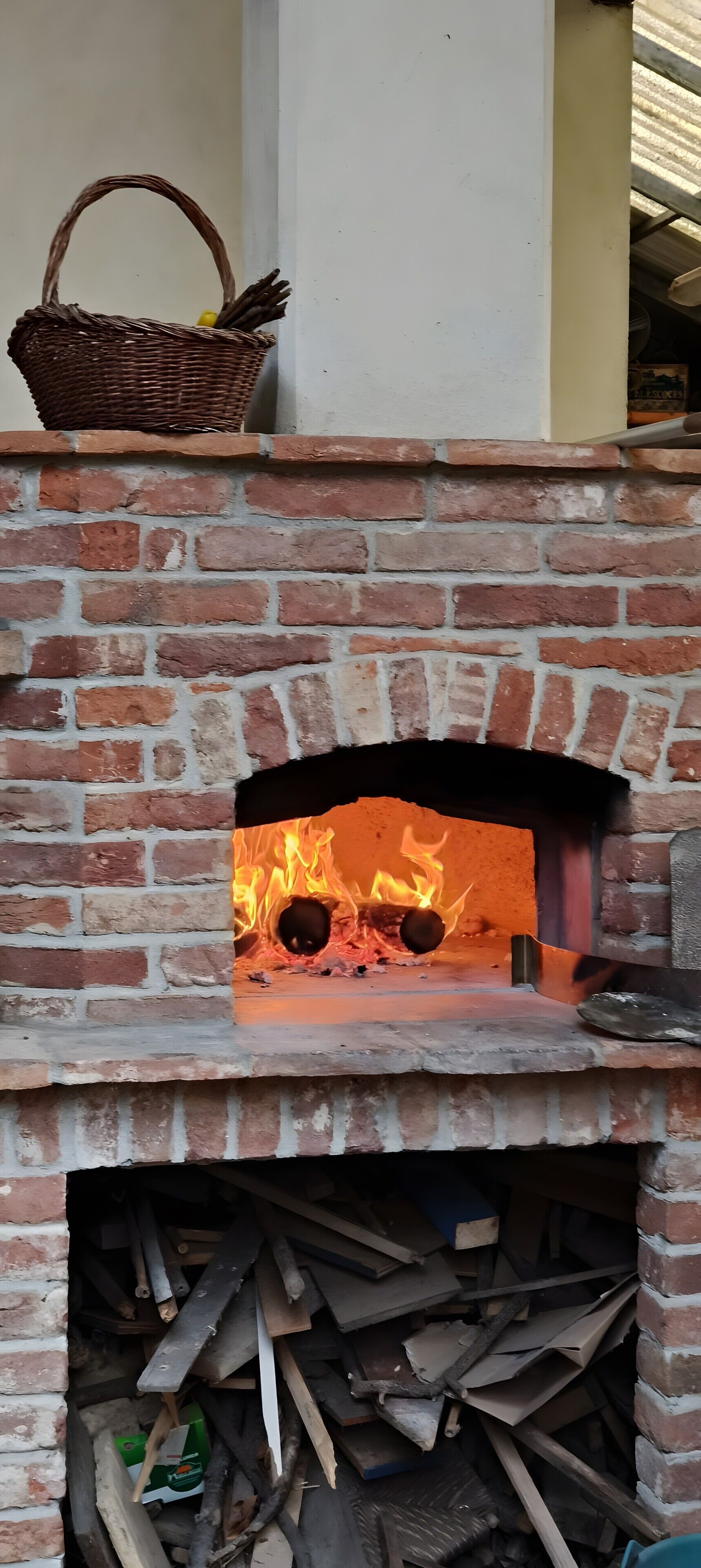 A brick fireplace with a fire burning inside, a basket on top, and a stack of firewood below.
