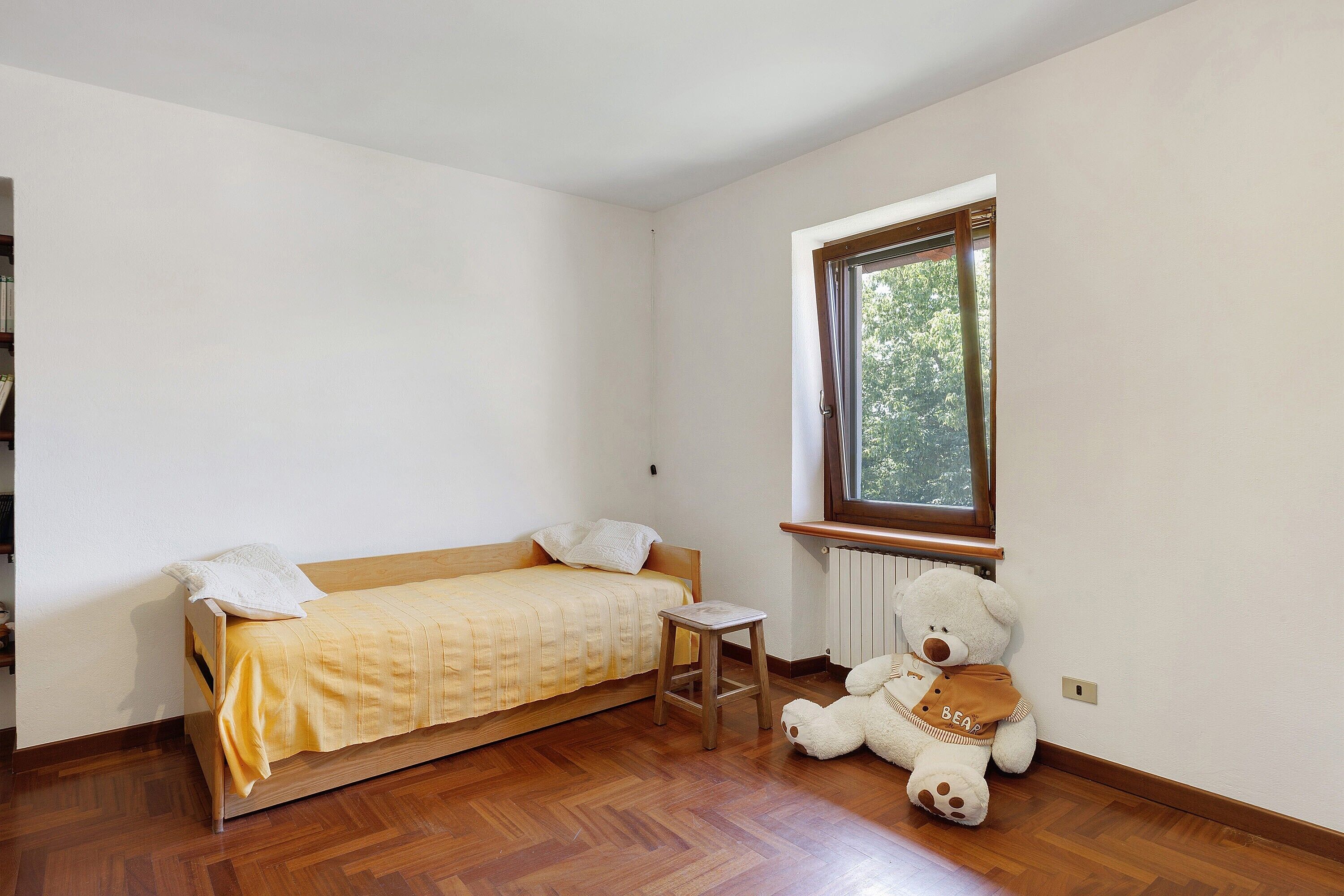 A single bed with a wooden frame and a yellow bedspread, a small wooden stool, and a teddy bear by a window.