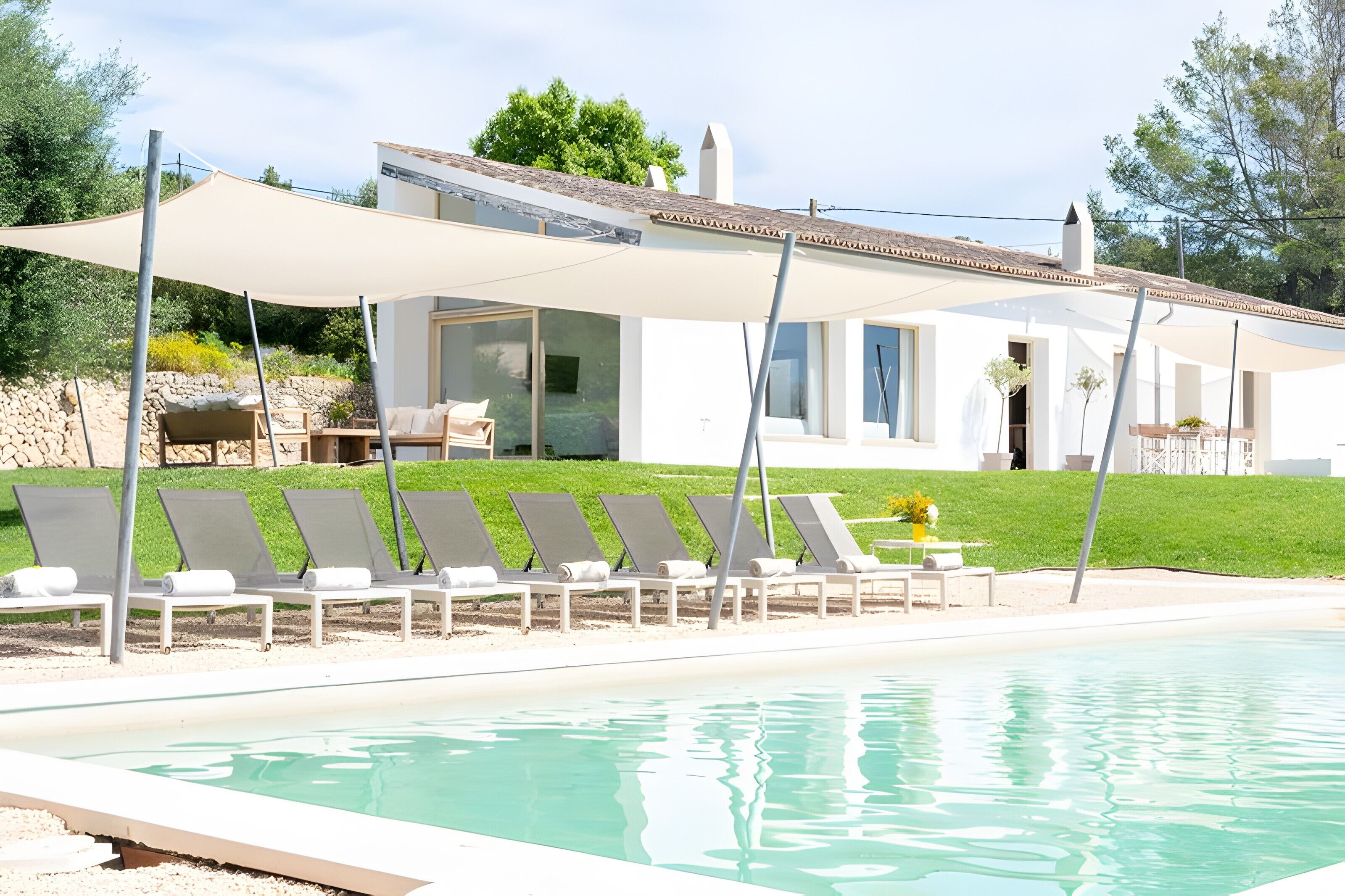 Zona de piscina con sillones de descanso y una pérgola, un edificio blanco con ventanales grandes y un muro de piedra al fondo.