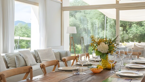 Un comedor con una mesa de madera dispuesta para cuatro personas, rodeado de sillas blancas, un sofá con cojines y un ventanal con vistas a zonas verdes.