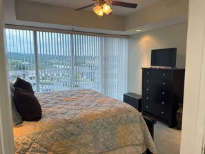 A bedroom with a bed, a dresser, a television, and a view of a cityscape through large windows.