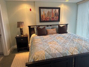 A neatly arranged bedroom with a bed, a nightstand, a lamp, a framed cityscape painting, and a window with blinds.