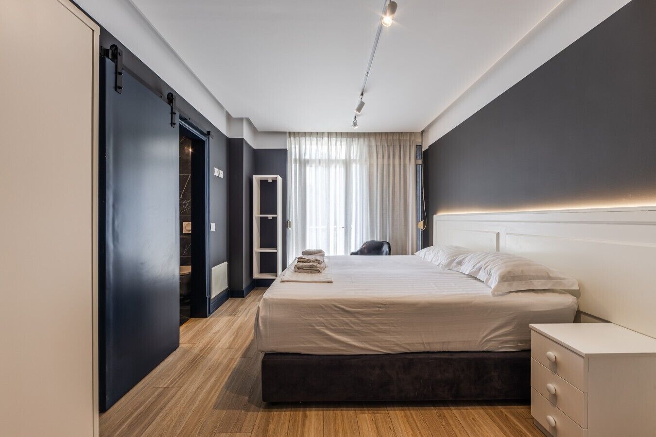A modern hotel room with a large bed, bedside tables, a wardrobe, and a bathroom visible through an open door.