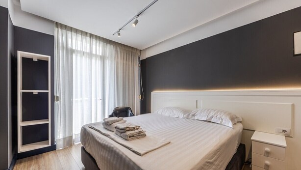 A modern hotel room with a large bed, a bedside table, a wardrobe, and a window with curtains.