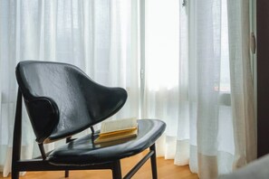 Una silla negra con un libro encima, ubicada cerca de una ventana con cortinas traslúcidas.