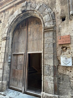 Un antiguo edificio de piedra con una gran puerta de madera y una escalera que conduce al interior. Hay dos placas en la pared, una que indica la importancia histórica del edificio y otra para un negocio.