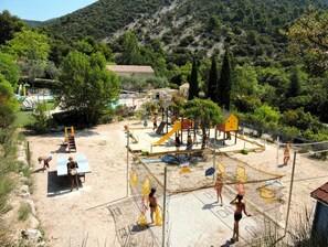 Ein Spielplatz mit verschiedenen Spielgeräten, darunter Rutschen und Kletterbereiche, umgeben von Bäumen und einer hügeligen Landschaft.