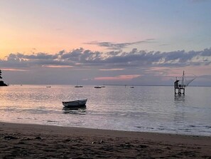 A beach with boats, a fishing net structure, and calm water during sunset.