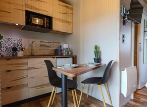 Une cuisine compacte avec des meubles en bois, un four à micro-ondes, une machine à café et un réfrigérateur. On y trouve une table à manger avec deux chaises et un cactus en pot.