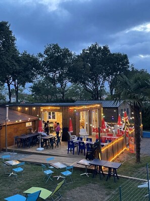 Un espace extĂ©rieur amĂ©nagĂ© pour les repas, avec des tables et des chaises, une terrasse en bois et un bĂątiment dotĂ© dâune porte en verre.