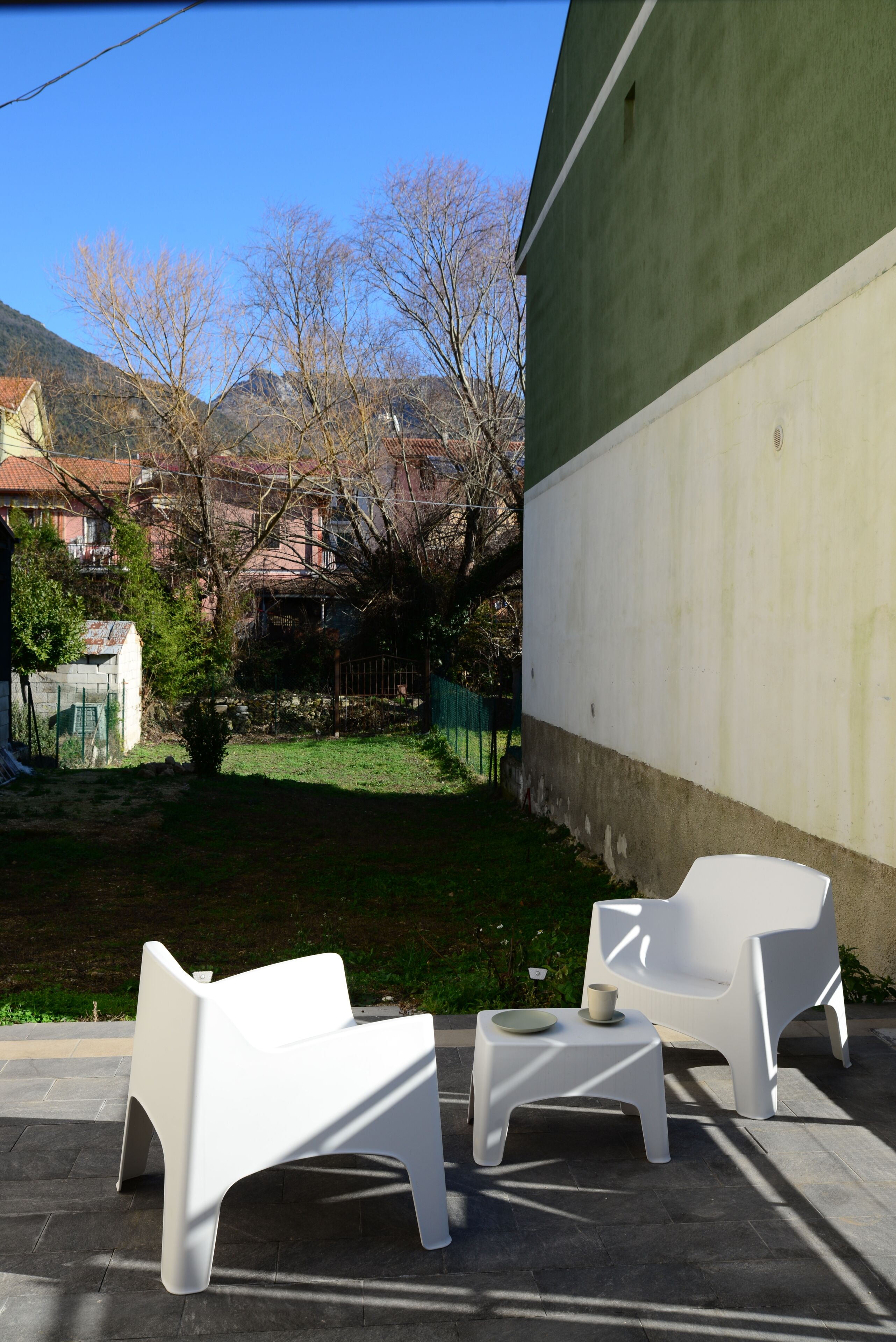 An outdoor seating area with two white chairs and a small table, a green wall, and a view of a residential area with trees and houses.