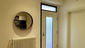 Un couloir avec un radiateur, un miroir rond, une porte dotée d’un panneau en verre dépoli et une porte blanche.